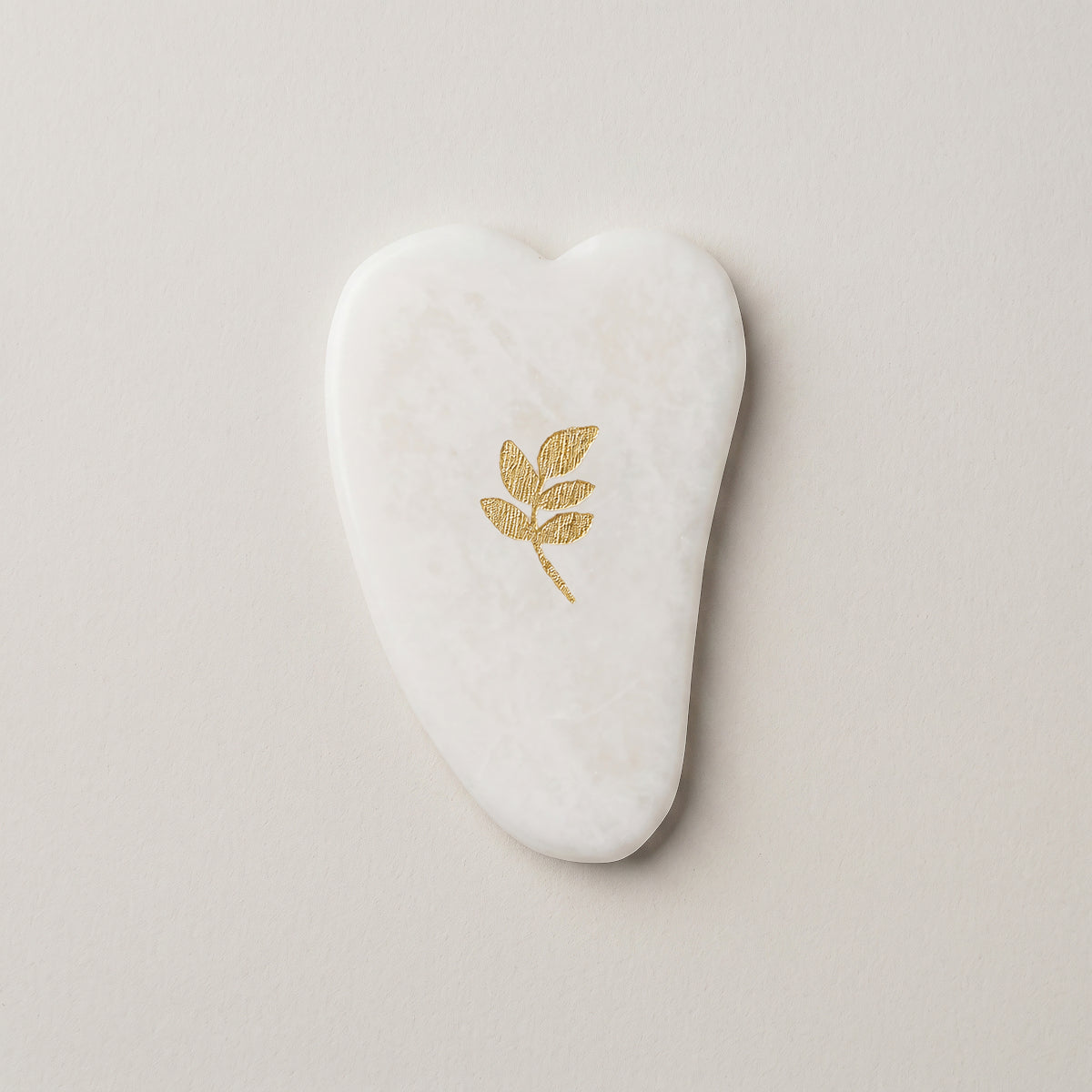 Gua Sha SCULPTIFY din jad alb cu vene fine, în formă de inimă alungită, cu motiv auriu de frunze, așezat plat, văzut de sus
