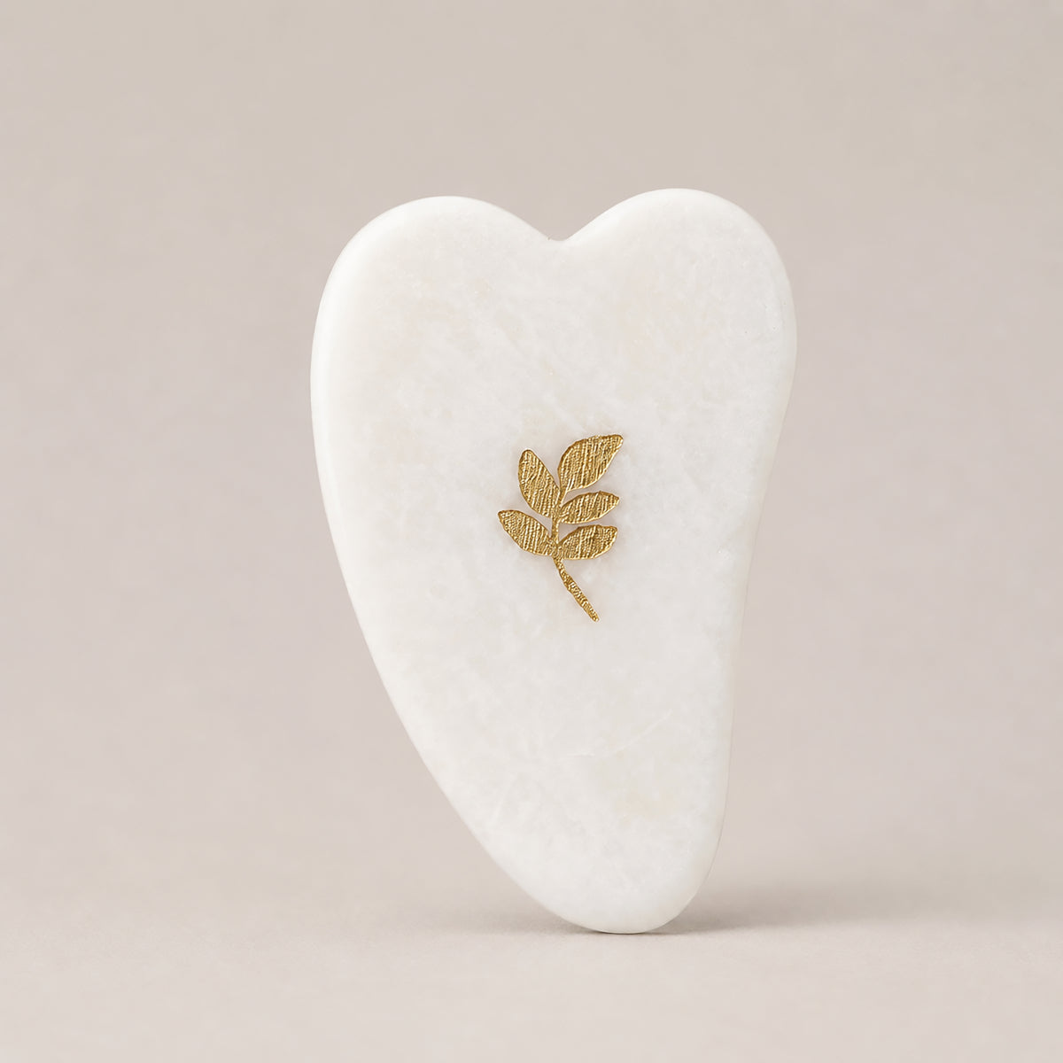 Gua Sha SCULPTIFY pentru masaj facial, din jad alb cu vene fine, în formă de inimă alungită, cu motiv auriu, așezat vertical