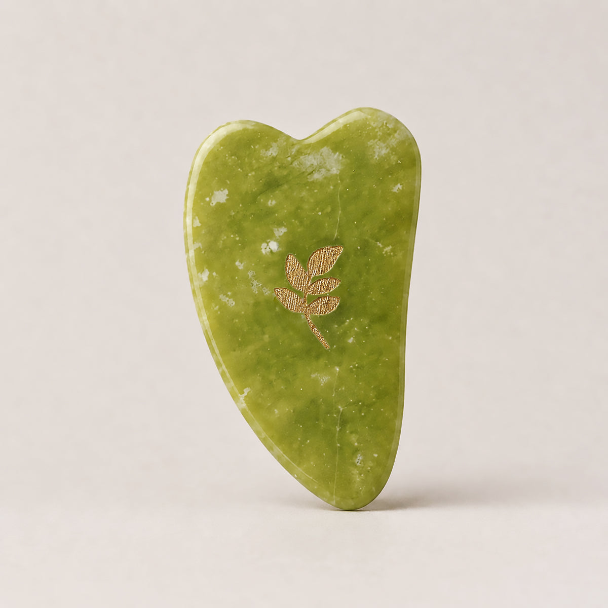 Gua Sha SCULPTIFY pentru masaj facial, din jad verde cu pete albe, în formă de inimă alungită, cu motiv auriu, așezat vertical
